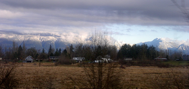 [The foreground is brown dried grassy vegetation with some trees. Behind the buildings is a row of evergreens and behind those are the mountains. The top of the mountain on the right is visible due to clear skies over it. The ones in the middle and the left are partially obscured by a blanet of clouds, but snow is visible on the lower levels of the mountains.]